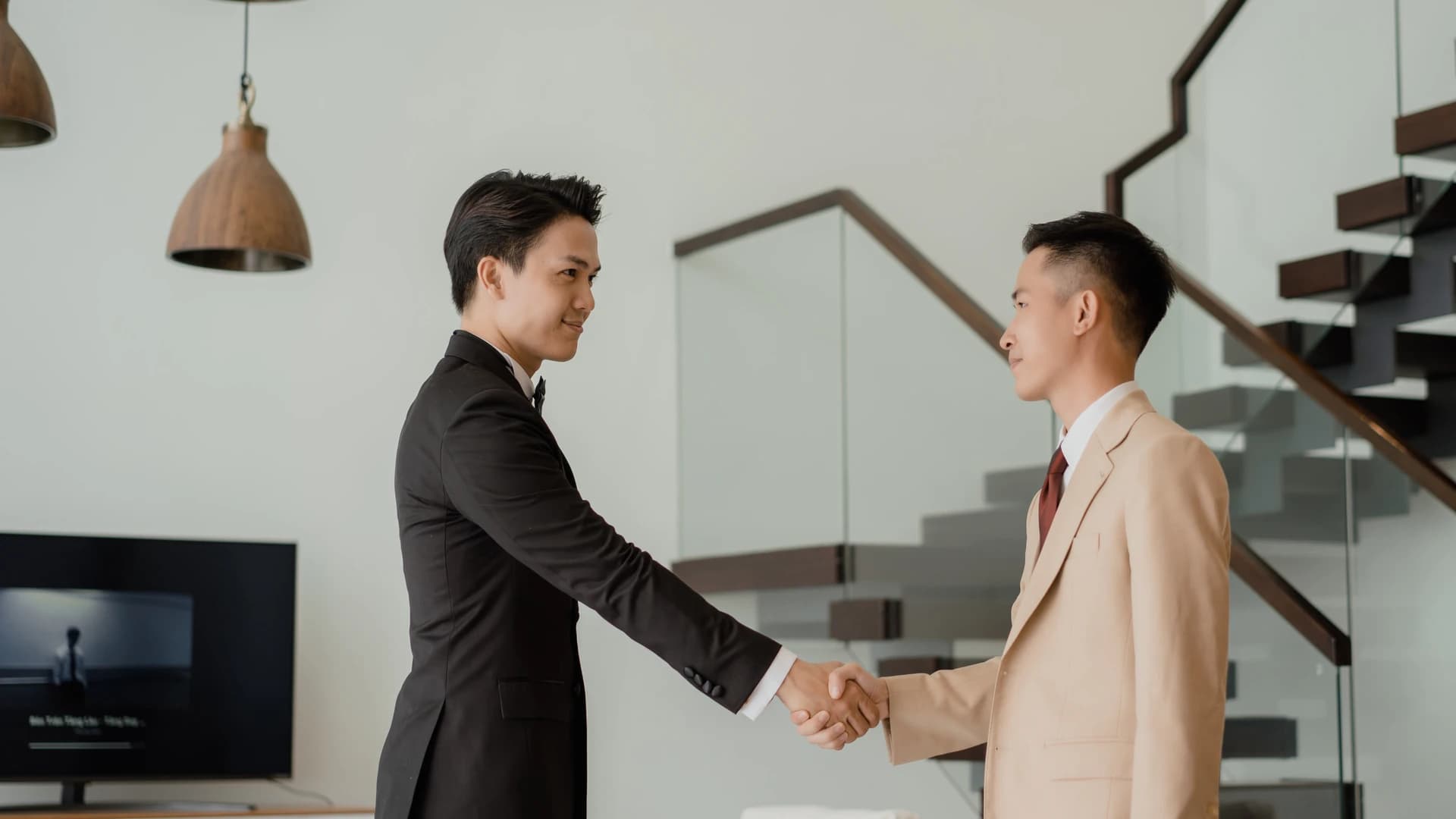 Hai người đàn ông bắt tay trong căn hộ sáng, một người mặc vest nâu, một người vest be