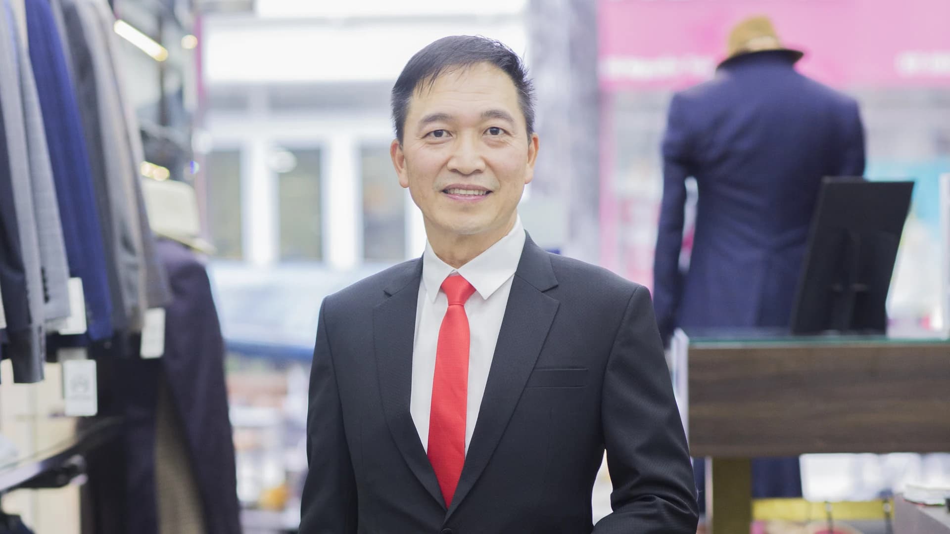 Quý ông tươi cười mặc vest đen, cà vạt đỏ tại showroom Mon Amie