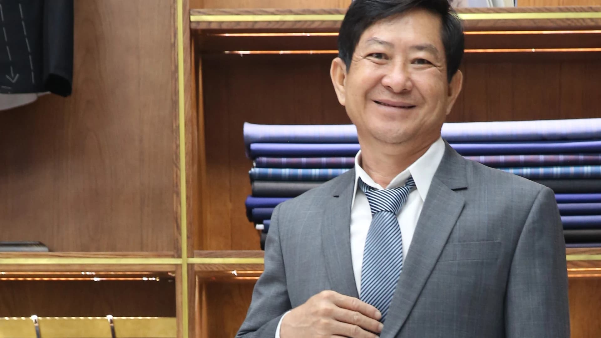 Quý ông cười tươi trong vest xám, cà vạt sọc, phía sau là kệ vải