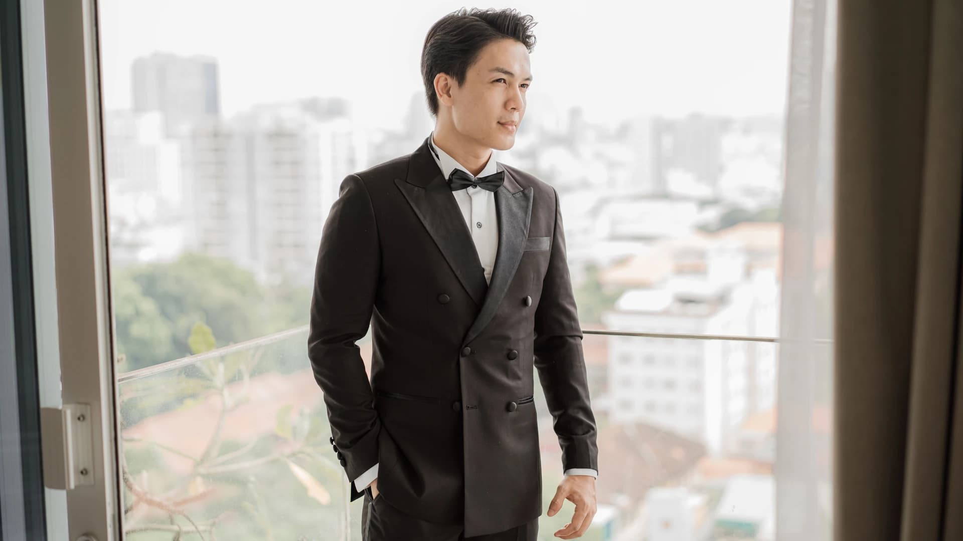 Chú rể mặc tuxedo đen cài nơ bên cửa sổ lớn nhìn ra thành phố