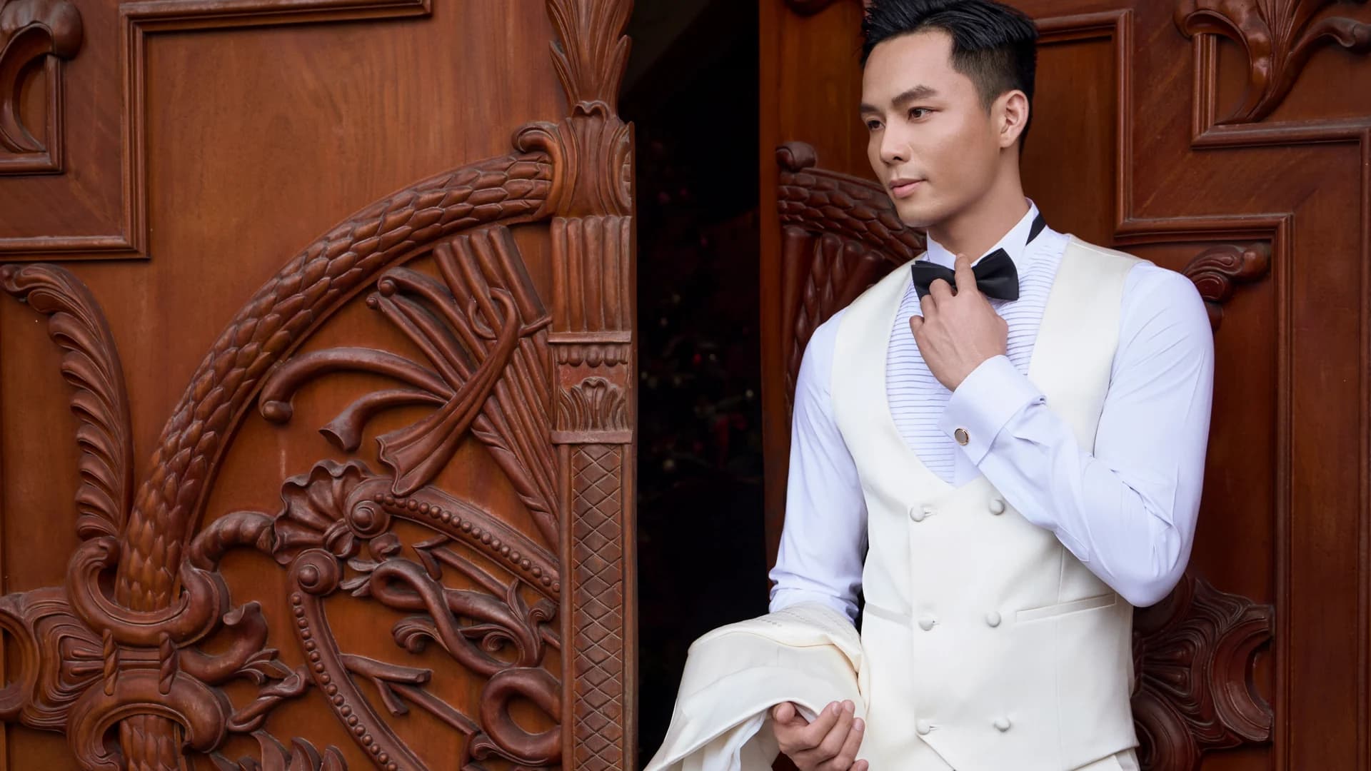 Groom in a cream double-breasted waistcoat and black bow tie beside a carved wooden door